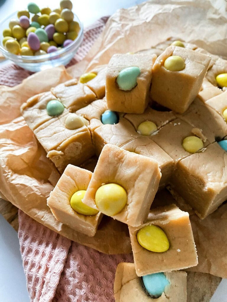 Easter Mini Eggs Fudge, Delicious And With No Condensed Milk 6 Mini eggs fudge chunks on parchment paper over a pink napkin and with mini chocolate eggs scattered around the marble countertop