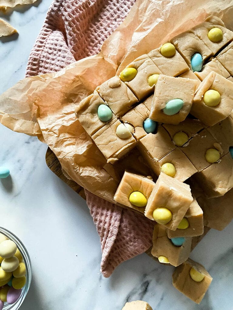 Easter Mini Eggs Fudge, Delicious And With No Condensed Milk 7 Mini eggs fudge chunks on parchment paper over a pink napkin and with mini chocolate eggs scattered around the marble countertop