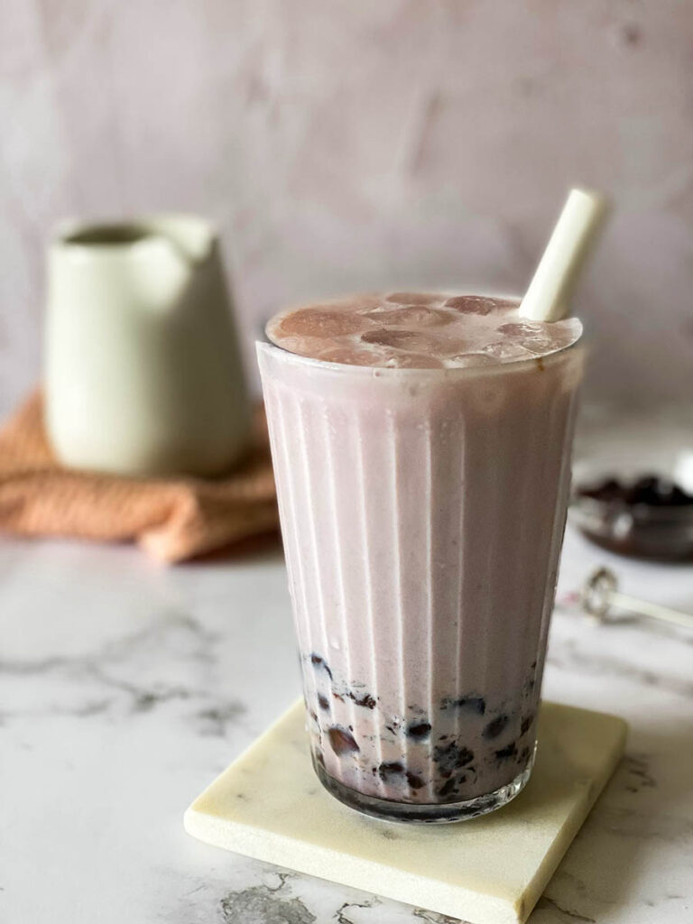 taro latte in a glass with a straw, on a marble coaster, jug, towel and boba pearls in the background, on a marble background