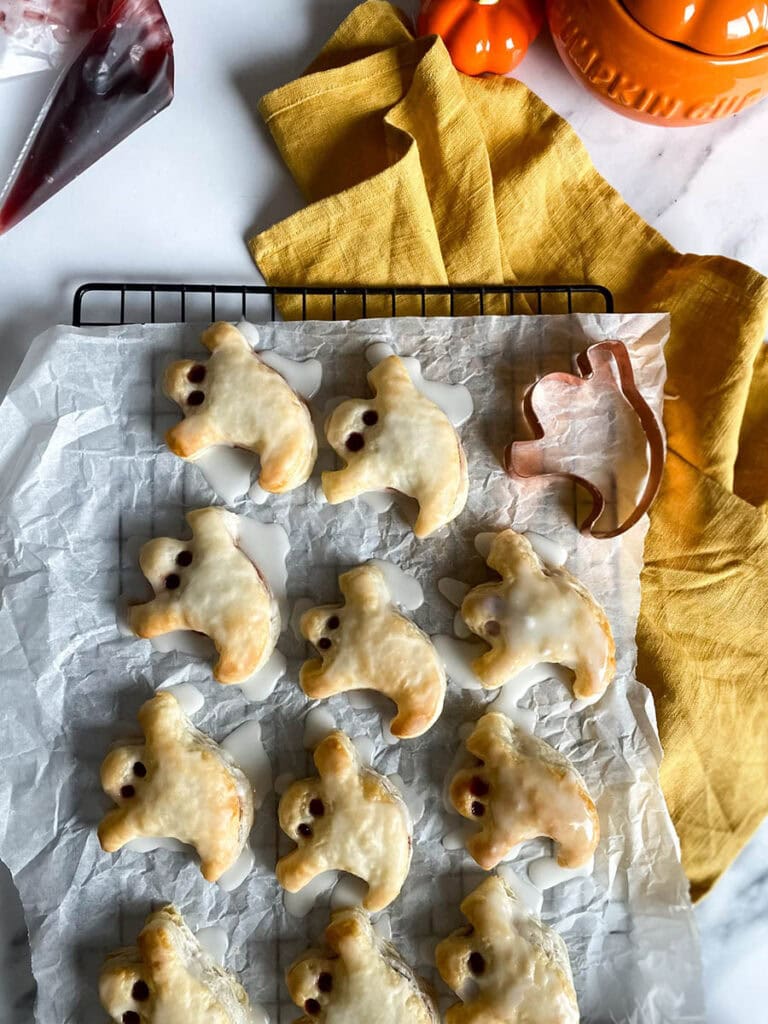 Ghost Hand Pies: Cute & Delicious Halloween Dessert 2 A tray of ghost hand pies is cooling on a wire rack covered with parchment paper. The pies are iced with a white glaze, and their "eyes" are made with small dark filling dots. A copper ghost-shaped cookie cutter and a yellow kitchen towel are placed nearby for decoration.
