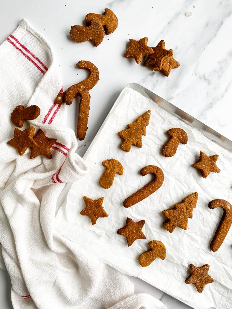 Easy Gingerbread Cookies For Dogs, 6 Ingredients Only 1 Homemade gingerbread cookies for dogs shaped like stars, candy canes, and Christmas trees, arranged on parchment paper and a white towel with red stripes. These rustic, wholesome treats make a festive, dog-friendly snack option for the holiday season.