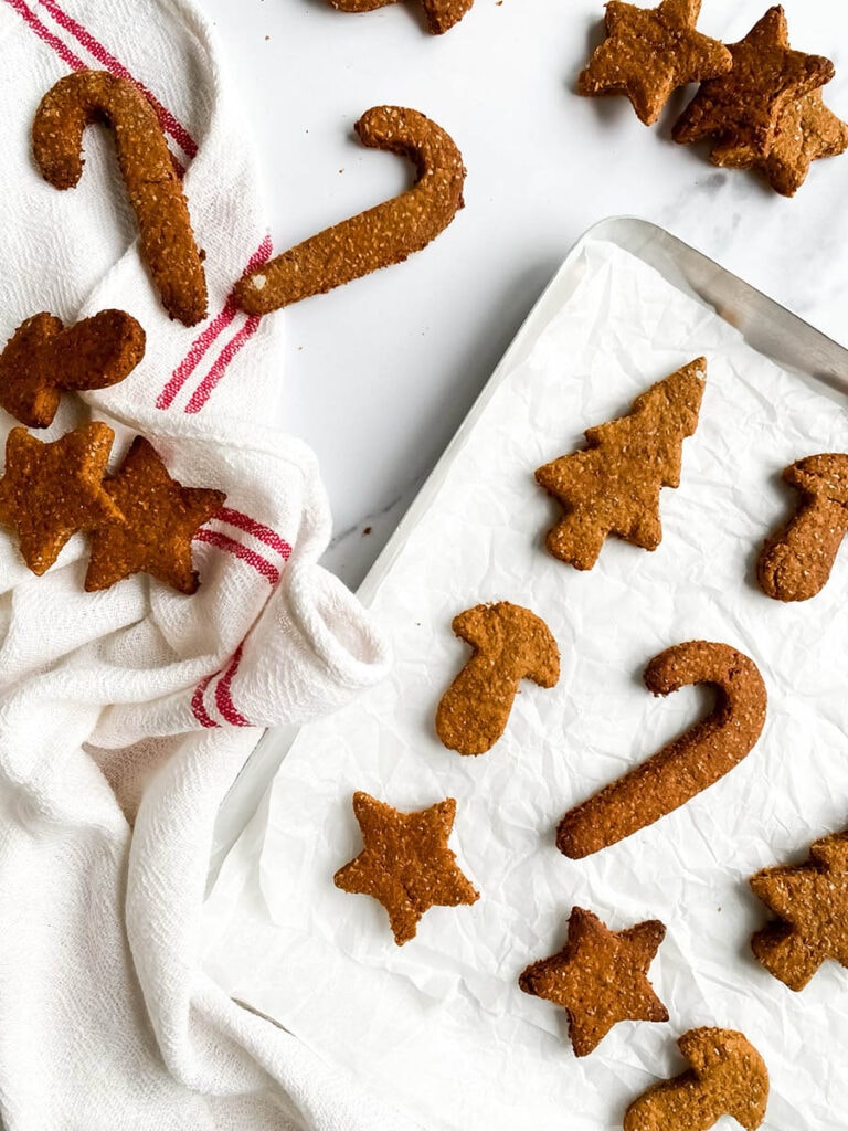 Easy Gingerbread Cookies For Dogs, 6 Ingredients Only 2 Homemade gingerbread cookies for dogs shaped like stars, candy canes, and Christmas trees, arranged on parchment paper and a white towel with red stripes. These rustic, wholesome treats make a festive, dog-friendly snack option for the holiday season.