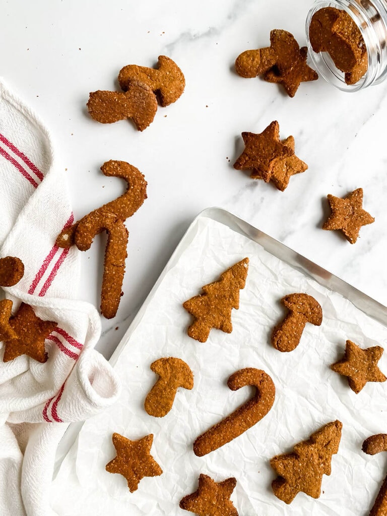 Easy Gingerbread Cookies For Dogs, 6 Ingredients Only 3 Homemade gingerbread cookies for dogs shaped like stars, candy canes, and Christmas trees, arranged on parchment paper and a white towel with red stripes. These rustic, wholesome treats make a festive, dog-friendly snack option for the holiday season.