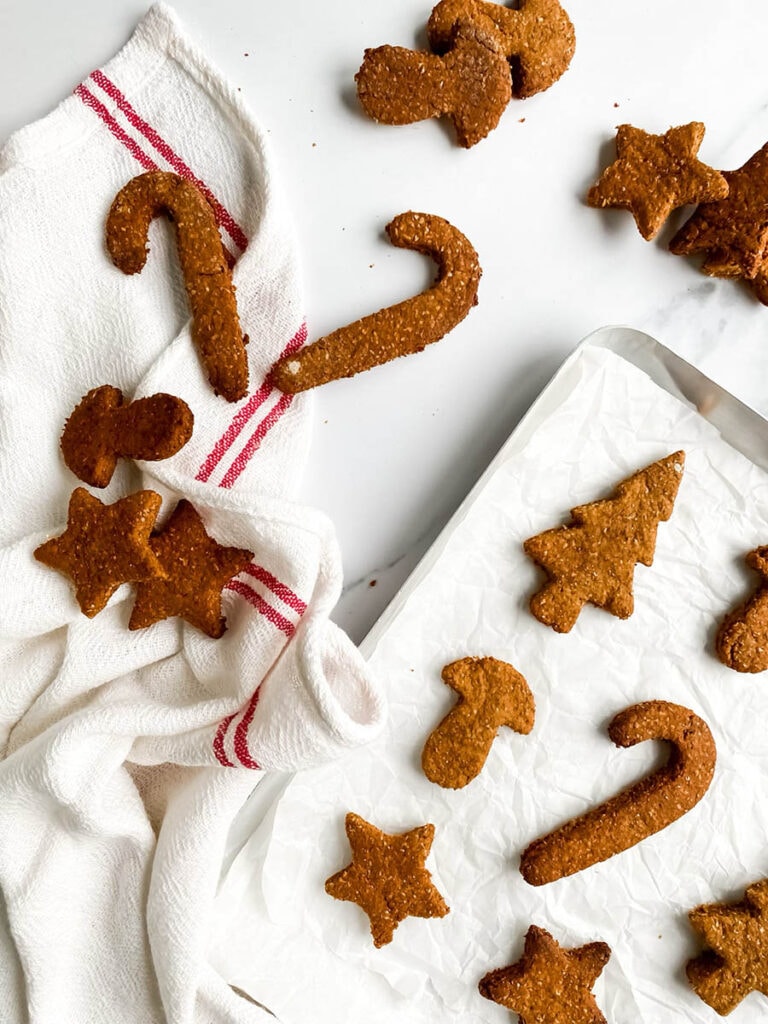 Easy Gingerbread Cookies For Dogs, 6 Ingredients Only 8 Homemade gingerbread cookies for dogs shaped like stars, candy canes, and Christmas trees, arranged on parchment paper and a white towel with red stripes. These rustic, wholesome treats make a festive, dog-friendly snack option for the holiday season.