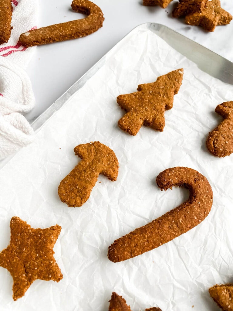 Easy Gingerbread Cookies For Dogs, 6 Ingredients Only 4 Homemade gingerbread cookies for dogs shaped like stars, candy canes, and Christmas trees, arranged on parchment paper and a white towel with red stripes. These rustic, wholesome treats make a festive, dog-friendly snack option for the holiday season.
