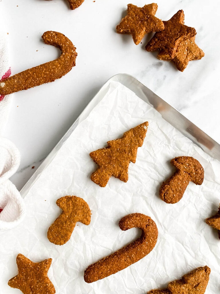 Easy Gingerbread Cookies For Dogs, 6 Ingredients Only 9 Homemade gingerbread cookies for dogs shaped like stars, candy canes, and Christmas trees, arranged on parchment paper and a white towel with red stripes. These rustic, wholesome treats make a festive, dog-friendly snack option for the holiday season.