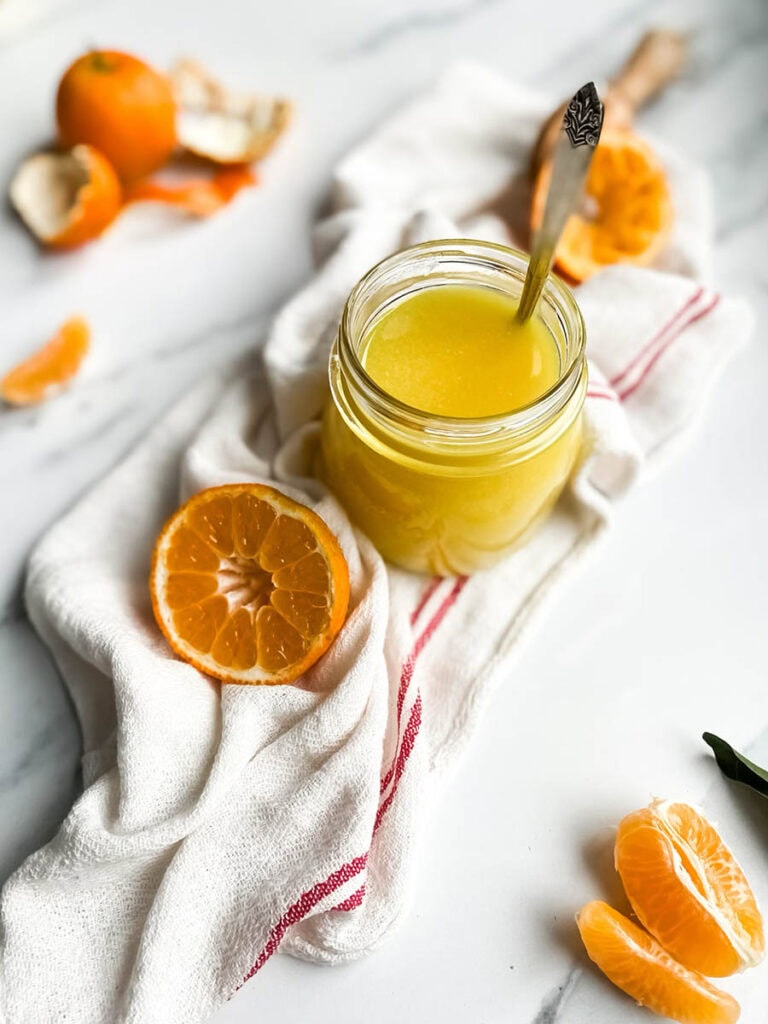 Easy Vegan Clementine Curd Recipe 3 A jar of bright orange clementine curd with a spoon, resting on a white cloth with red stripes. Fresh clementine slices and peeled segments are scattered around, along with a wooden citrus reamer, creating a fresh and vibrant setting. The background is a clean white marble surface, emphasizing the homemade citrus spread.
