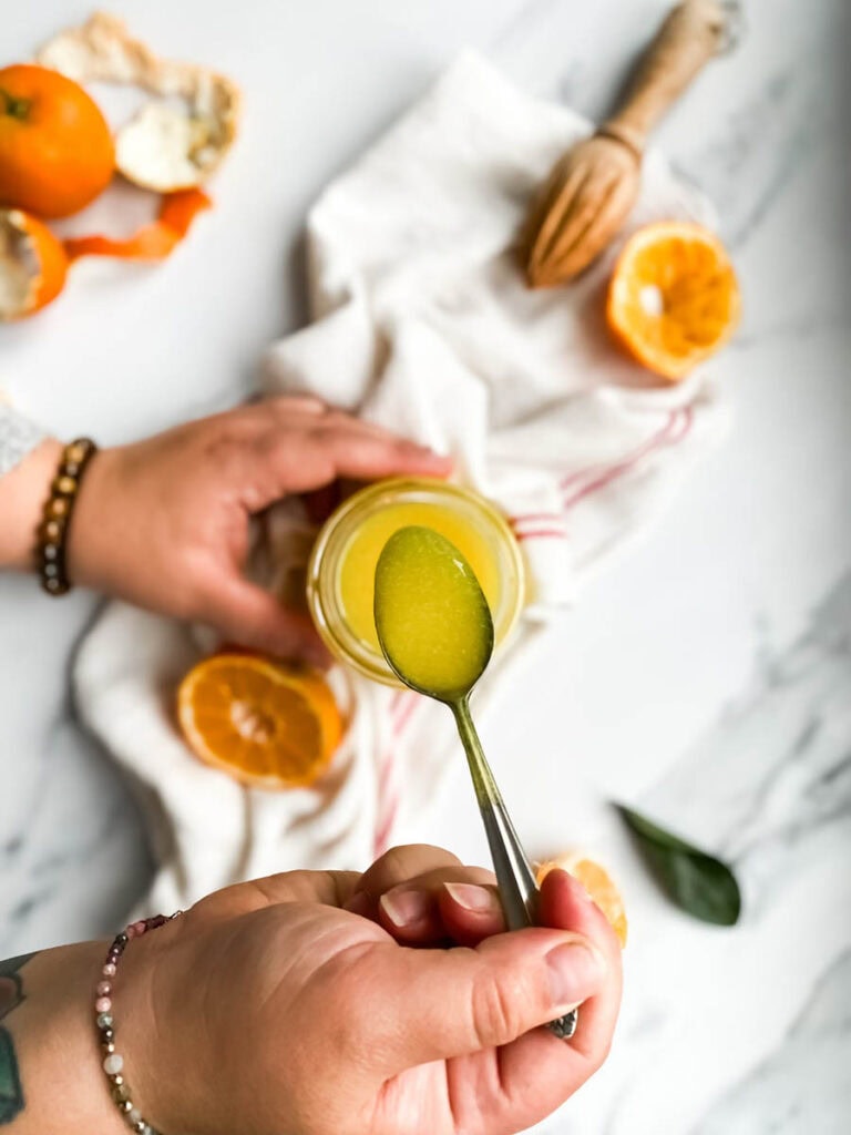 Easy Vegan Clementine Curd Recipe 4 A jar of bright orange clementine curd with a spoon, resting on a white cloth with red stripes. Fresh clementine slices and peeled segments are scattered around, along with a wooden citrus reamer, creating a fresh and vibrant setting. The background is a clean white marble surface, emphasizing the homemade citrus spread.