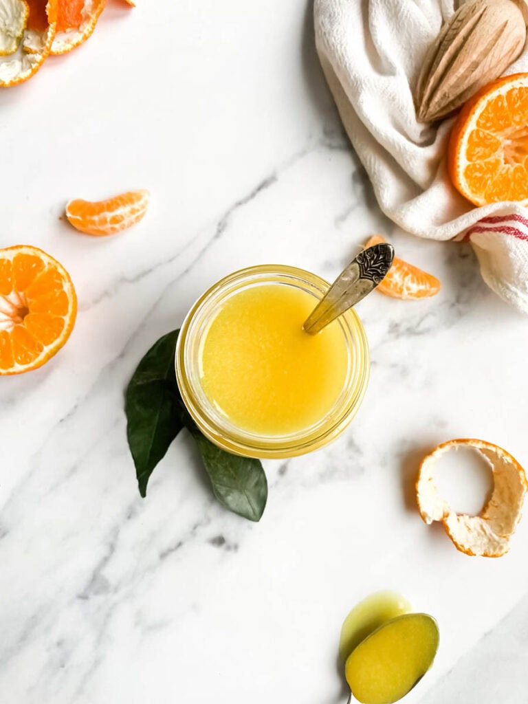 Easy Vegan Clementine Curd Recipe 5 A jar of bright orange clementine curd with a spoon, resting on a white cloth with red stripes. Fresh clementine slices and peeled segments are scattered around, along with a wooden citrus reamer, creating a fresh and vibrant setting. The background is a clean white marble surface, emphasizing the homemade citrus spread.