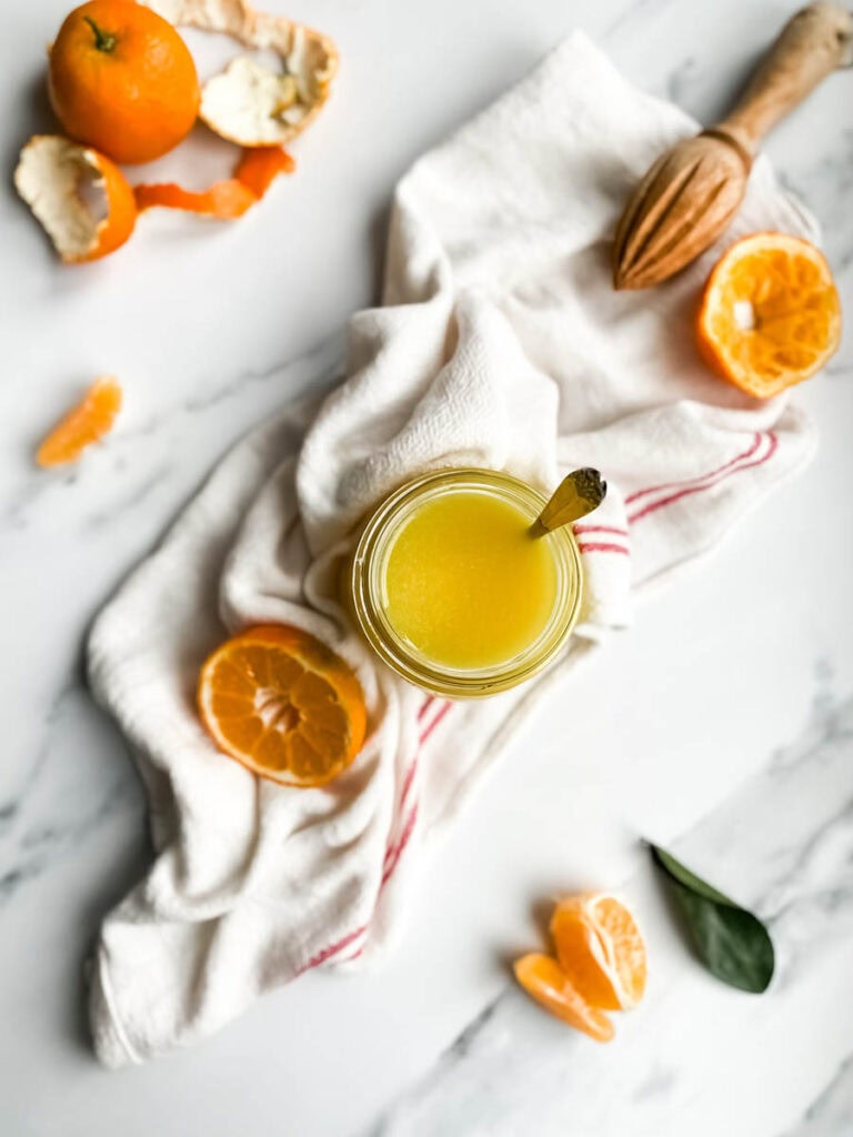 Easy Vegan Clementine Curd Recipe 8 A jar of bright orange clementine curd with a spoon, resting on a white cloth with red stripes. Fresh clementine slices and peeled segments are scattered around, along with a wooden citrus reamer, creating a fresh and vibrant setting. The background is a clean white marble surface, emphasizing the homemade citrus spread.