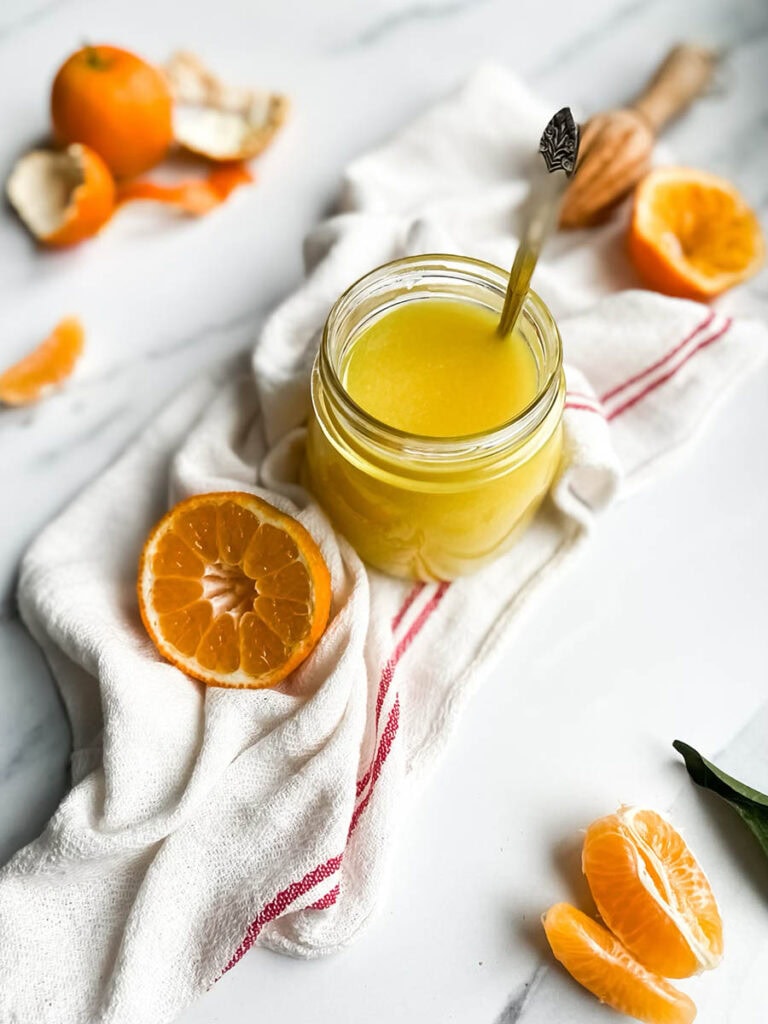 Easy Vegan Clementine Curd Recipe 9 A jar of bright orange clementine curd with a spoon, resting on a white cloth with red stripes. Fresh clementine slices and peeled segments are scattered around, along with a wooden citrus reamer, creating a fresh and vibrant setting. The background is a clean white marble surface, emphasizing the homemade citrus spread.
