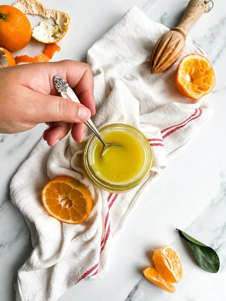 Easy Vegan Clementine Curd Recipe 10 A jar of bright orange clementine curd with a spoon, resting on a white cloth with red stripes. Fresh clementine slices and peeled segments are scattered around, along with a wooden citrus reamer, creating a fresh and vibrant setting. The background is a clean white marble surface, emphasizing the homemade citrus spread.