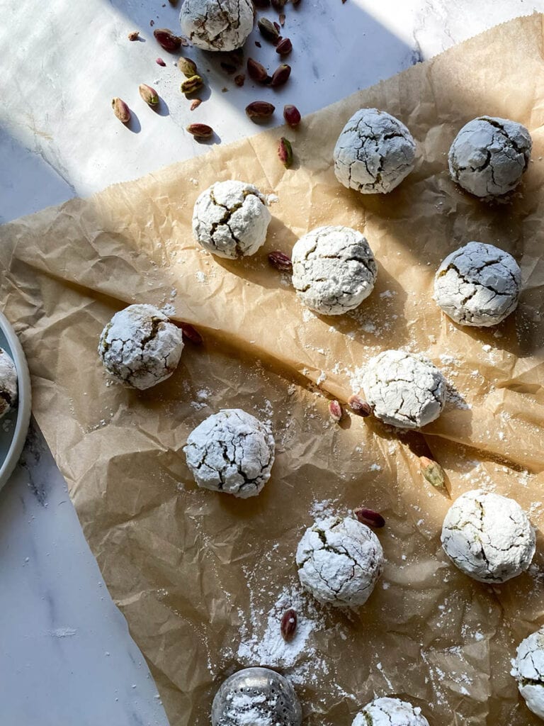 Yummy Pistachio Amaretti Cookies, For The Most Perfect Christmas 10 Multiple pistachio amaretti cookies, dusted with powdered sugar, are arranged on parchment paper with a few whole pistachios scattered around. The image showcases the rustic, crinkled texture of the cookies.