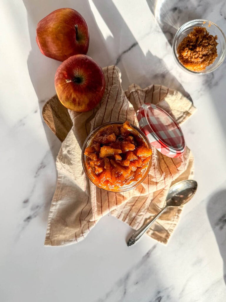 apples caramelized in a jar, over a striped towel, a spoon, apples and brown sugar on the sides, over a marble background