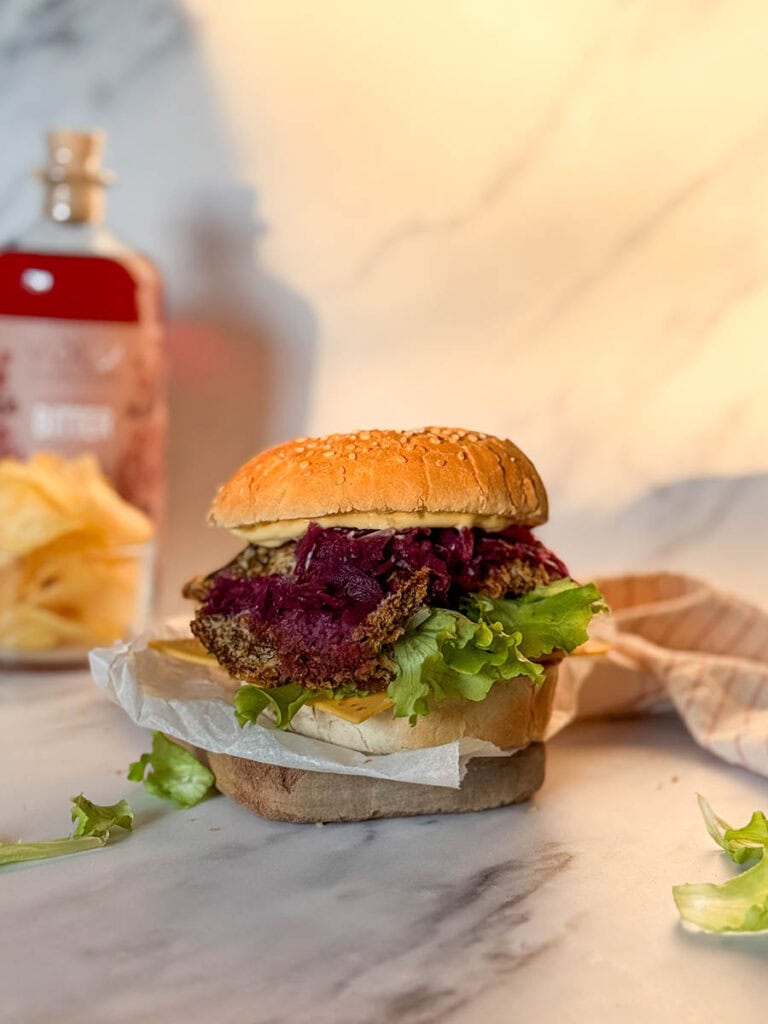 A golden, crispy vegan oyster mushroom burger stacked with fresh lettuce, cheese, and purple coleslaw, resting on parchment paper on a marble surface, surrounded by soft, natural light and a blurred background of condiments and chips.