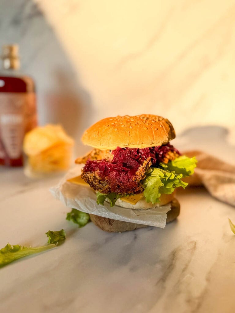A golden, crispy vegan oyster mushroom burger stacked with fresh lettuce, cheese, and purple coleslaw, resting on parchment paper on a marble surface, surrounded by soft, natural light and a blurred background of condiments and chips.