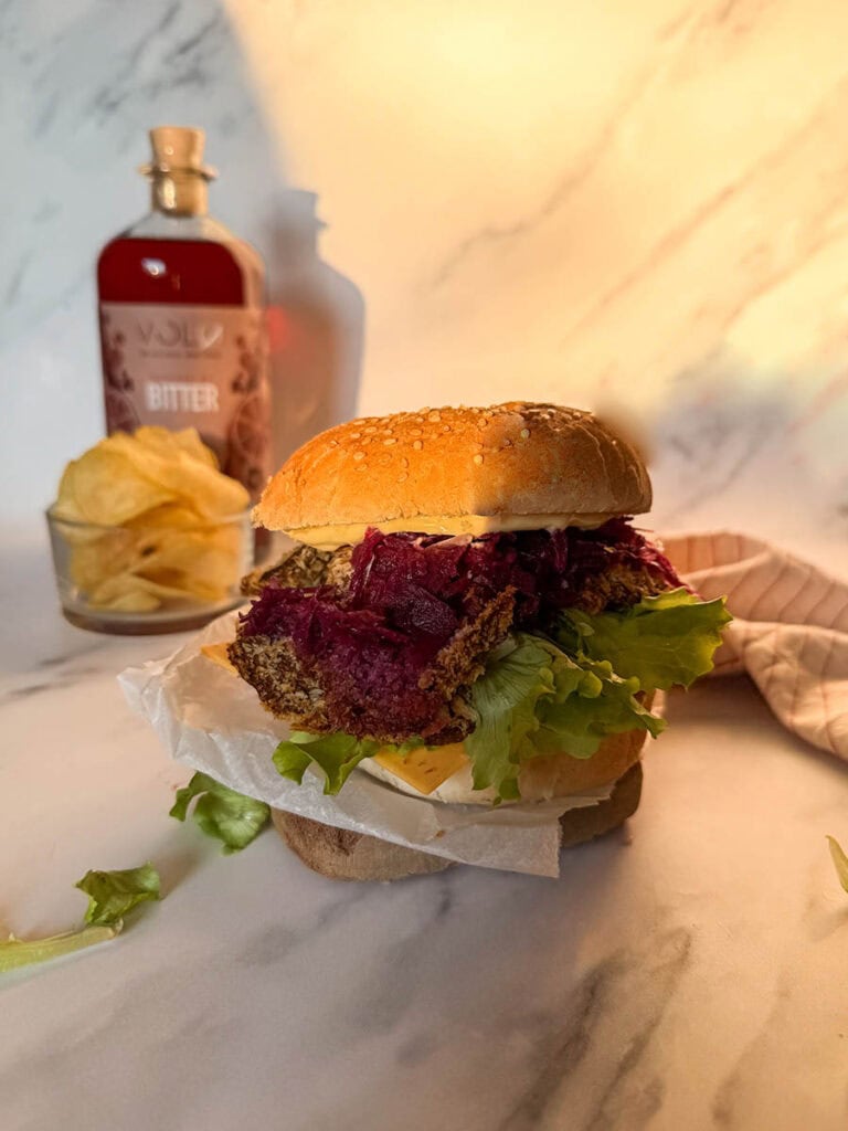 A close-up view of vegan oyster mushroom burgers featuring a crispy breaded mushroom patty, vibrant purple coleslaw, fresh lettuce, and cheese layered on a sesame seed bun, with a blurred background including chips and a sauce bottle.