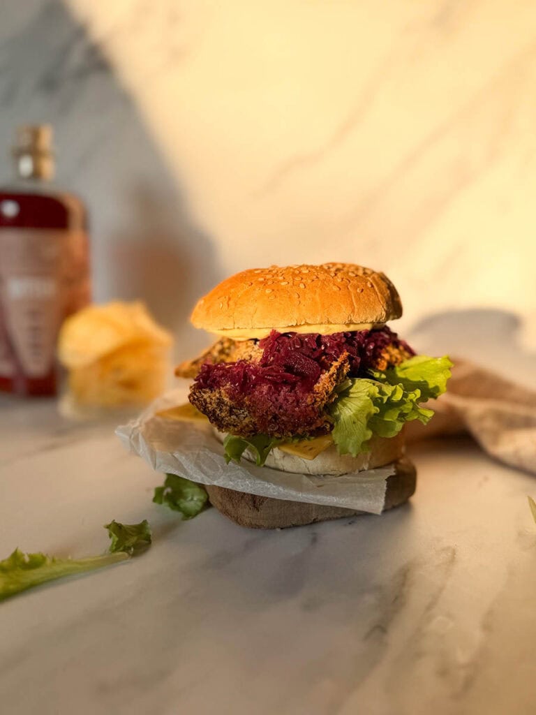 A golden, crispy vegan oyster mushroom burgers stacked with fresh lettuce, cheese, and purple coleslaw, resting on parchment paper on a marble surface, surrounded by soft, natural light and a blurred background of condiments and chips.