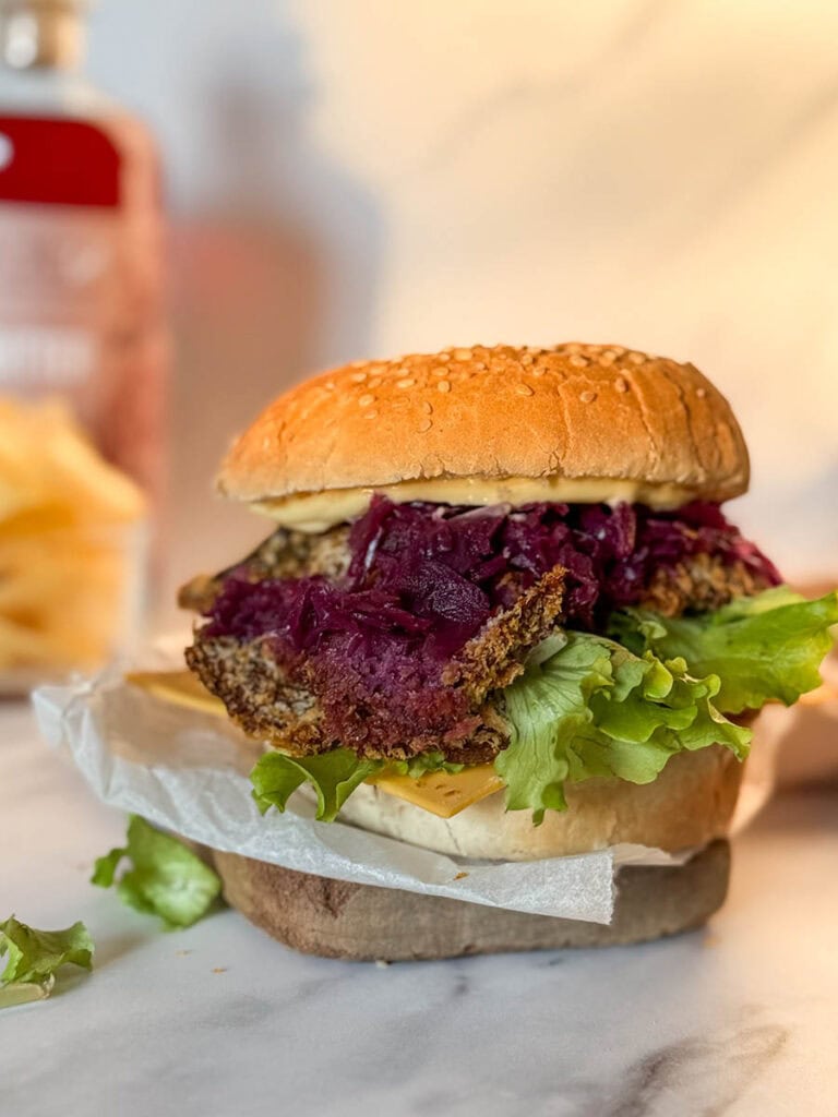 A close-up view of a vegan oyster mushroom burger featuring a crispy breaded mushroom patty, vibrant purple coleslaw, fresh lettuce, and cheese layered on a sesame seed bun, with a blurred background including chips and a sauce bottle.
