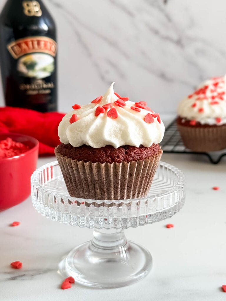 Baileys Red Velvet Cupcakes, The Perfect Valentine's Dessert 9 A Baileys red velvet cupcake with creamy frosting and red heart sprinkles on a glass stand, surrounded by scattered sprinkles, a Baileys bottle, and other cupcakes in the background.