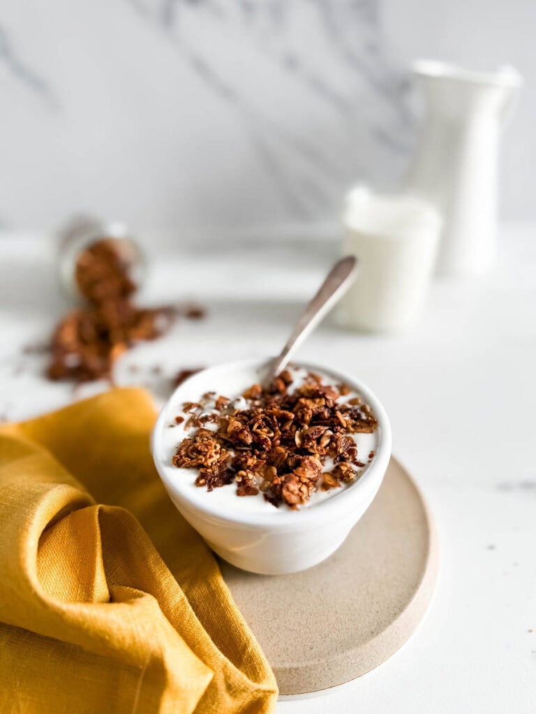 Granola with cinnamon in a bowl of creamy yogurt, placed on a simple beige plate. A vibrant yellow cloth and spilled granola enhance the cozy breakfast vibe, with a milk jar in the background.
