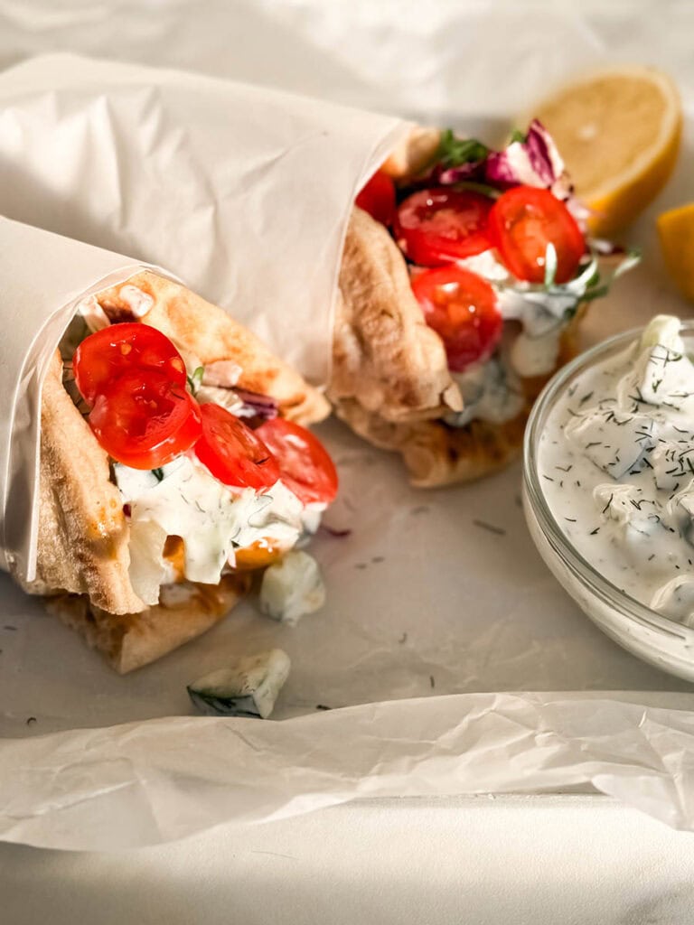 Simple Chicken Pitas, The Greek Way: Delicious And Quick 2 Close-up of chicken pitas, wrapped in parchment, highlighting juicy grilled chicken, bright red cherry tomatoes, and fresh greens. A small bowl of tzatziki with dill and cucumber chunks sits nearby.