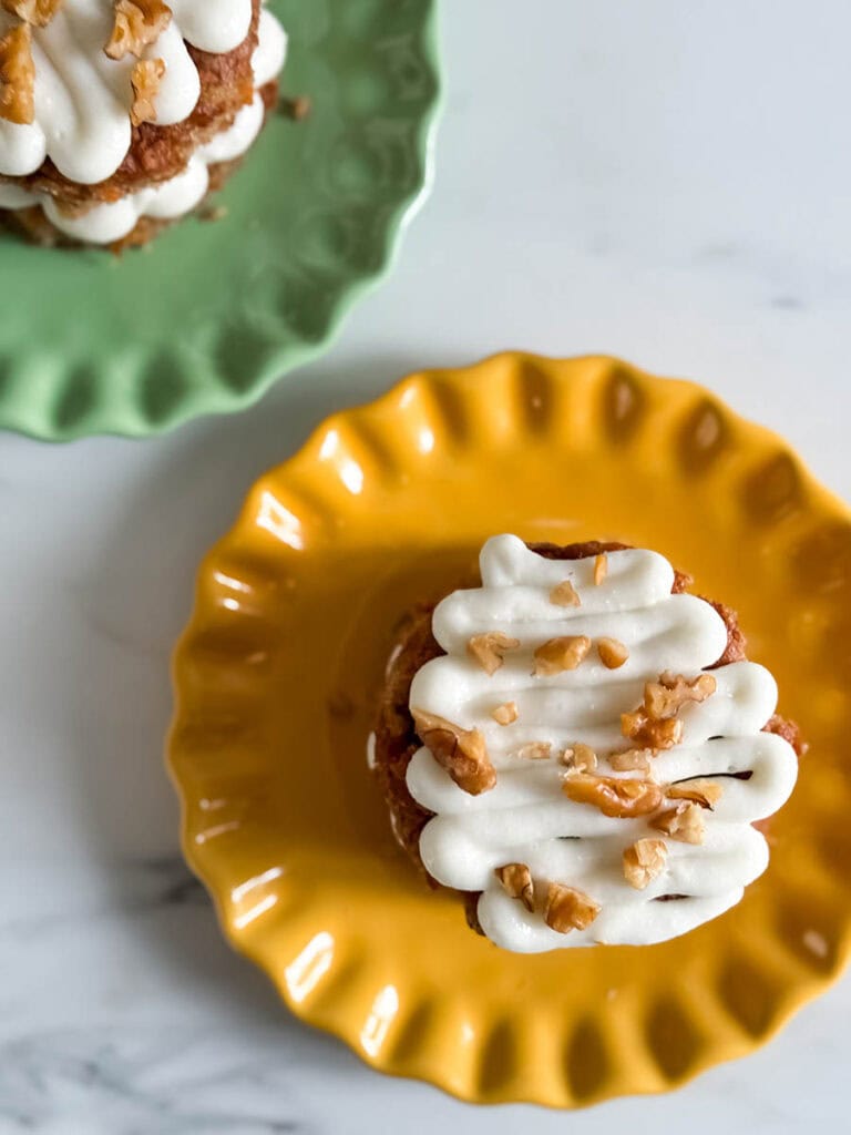 Mini Carrot Cake Recipe, The Perfect Easter Dessert 2 Top-down view of a mini carrot cake on a yellow scalloped plate, drizzled with piped cream cheese frosting and topped with chopped walnuts. A second cake is partially visible on a green plate in the background.