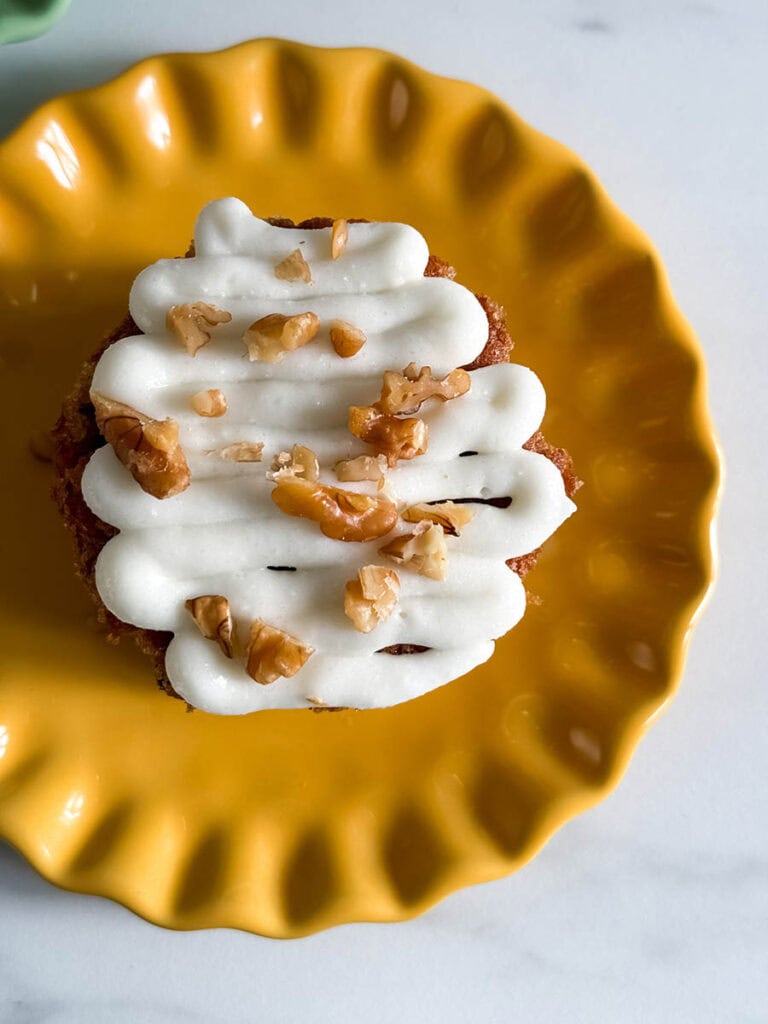 Mini Carrot Cake Recipe, The Perfect Easter Dessert 4 Close-up of a mini carrot cake with thick cream cheese frosting in a zig-zag pattern, sprinkled with walnuts, sitting on a bright yellow plate.