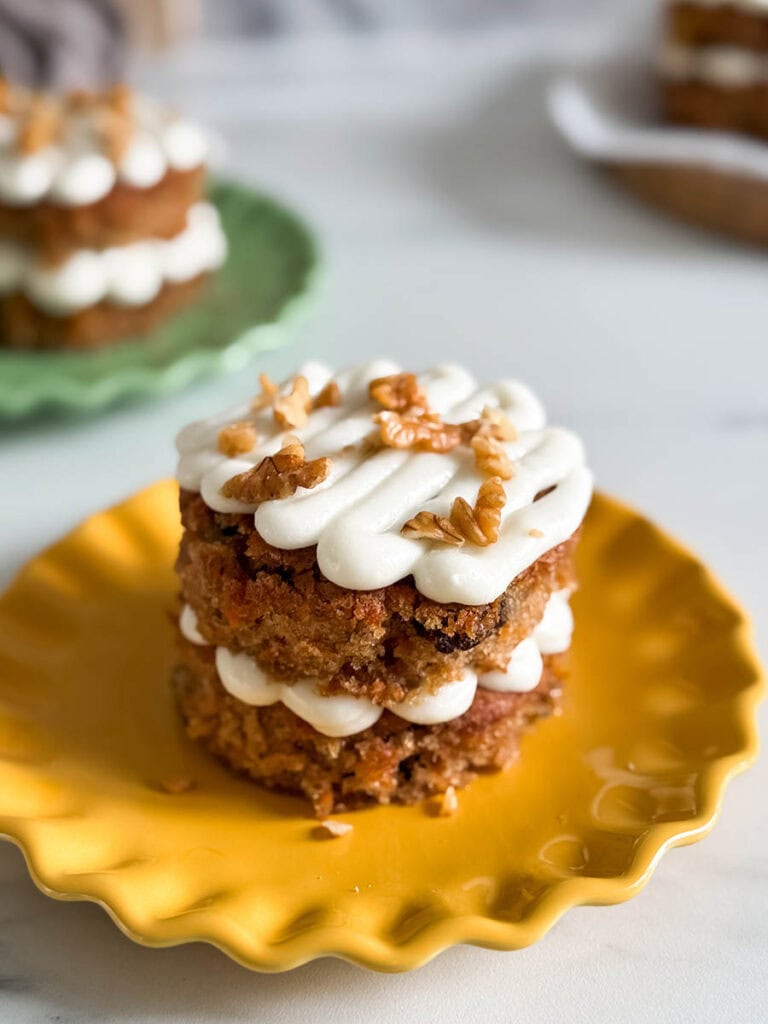 Mini Carrot Cake Recipe, The Perfect Easter Dessert 1 Another angle of a stacked carrot cake with creamy frosting and walnuts, placed on a yellow plate, with additional cakes blurred in the background.