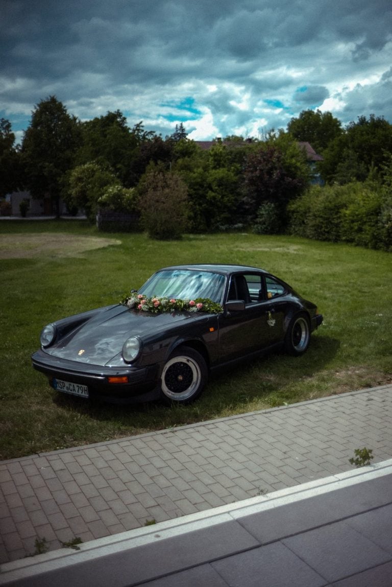 Ein schwarzer Porsche 911 Oldtimer mit Blumenschmuck auf der Motorhaube steht auf einer grünen Wiese vor einem bewaldeten Hintergrund bei Wolken.