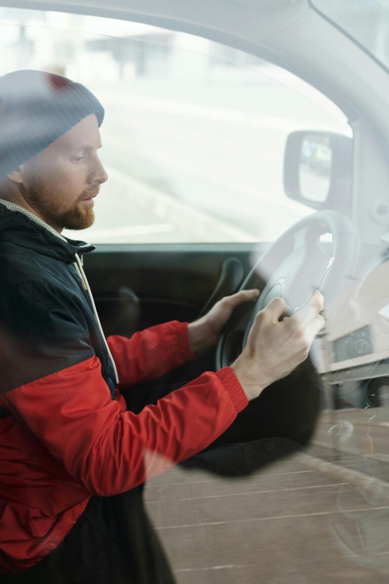 A man with a beard wearing a red jacket uses a smartphone while driving a car.