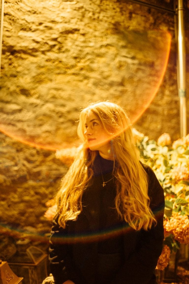 Eine junge Frau mit langem, hellem Haar steht vor einer Steinmauer, beleuchtet von warmem, goldenem Licht mit einem deutlichen Regenbogeneffekt.