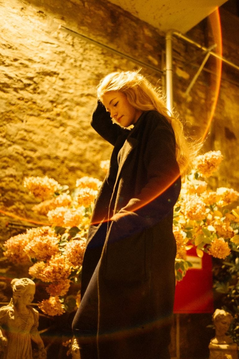 Eine junge Frau mit langem blonden Haar trägt einen dunklen Mantel und steht vor einer Steinmauer mit gelb beleuchteten Blumen und Statuen.