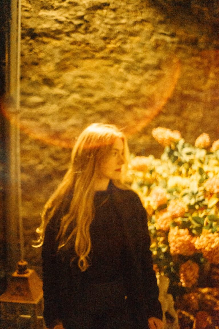 Eine junge Frau mit langem blondem Haar steht vor einer Steinmauer und Blumen in warmem, goldenem Licht mit einem leichten Lichteffekt im Bild.
