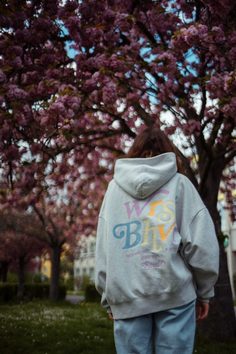 Eine Person von hinten in einem grauen Kapuzenpullover mit buntem Schriftzug vor einem blühenden Kirschbaum mit leuchtend rosa Blüten im Frühling.