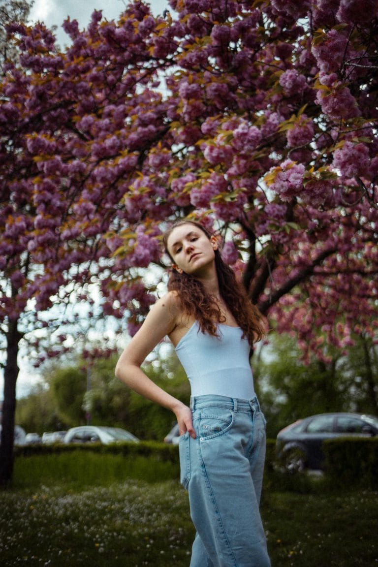 Eine junge Frau mit langen braunen Haaren steht in einem hellblauen Top und Jeans vor einem blühenden Kirschbaum mit leuchtend rosa Blüten.