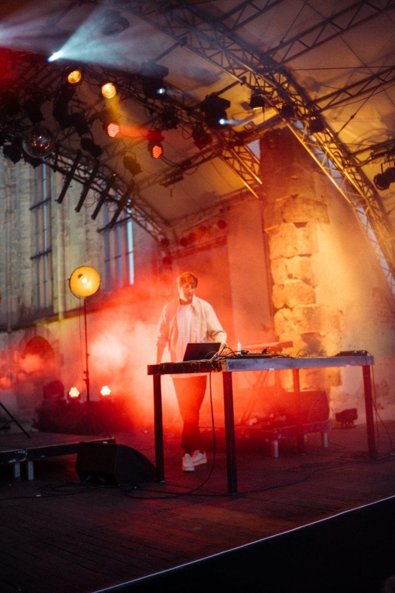 Musiker Christian Löffler steht an einem Pult auf einer Bühne, umgeben von rotem Bühnenlicht und dichtem Nebel in einer historischen Kulisse mit alten Steinmauern.