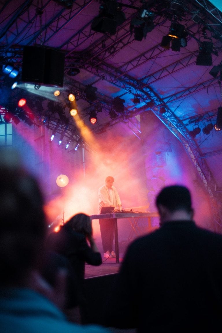 Ein DJ steht auf einer Bühne vor einem Mischpult, umgeben von dramatischem rotem und blauem Licht sowie Nebel bei einem Live Konzert am Abend.