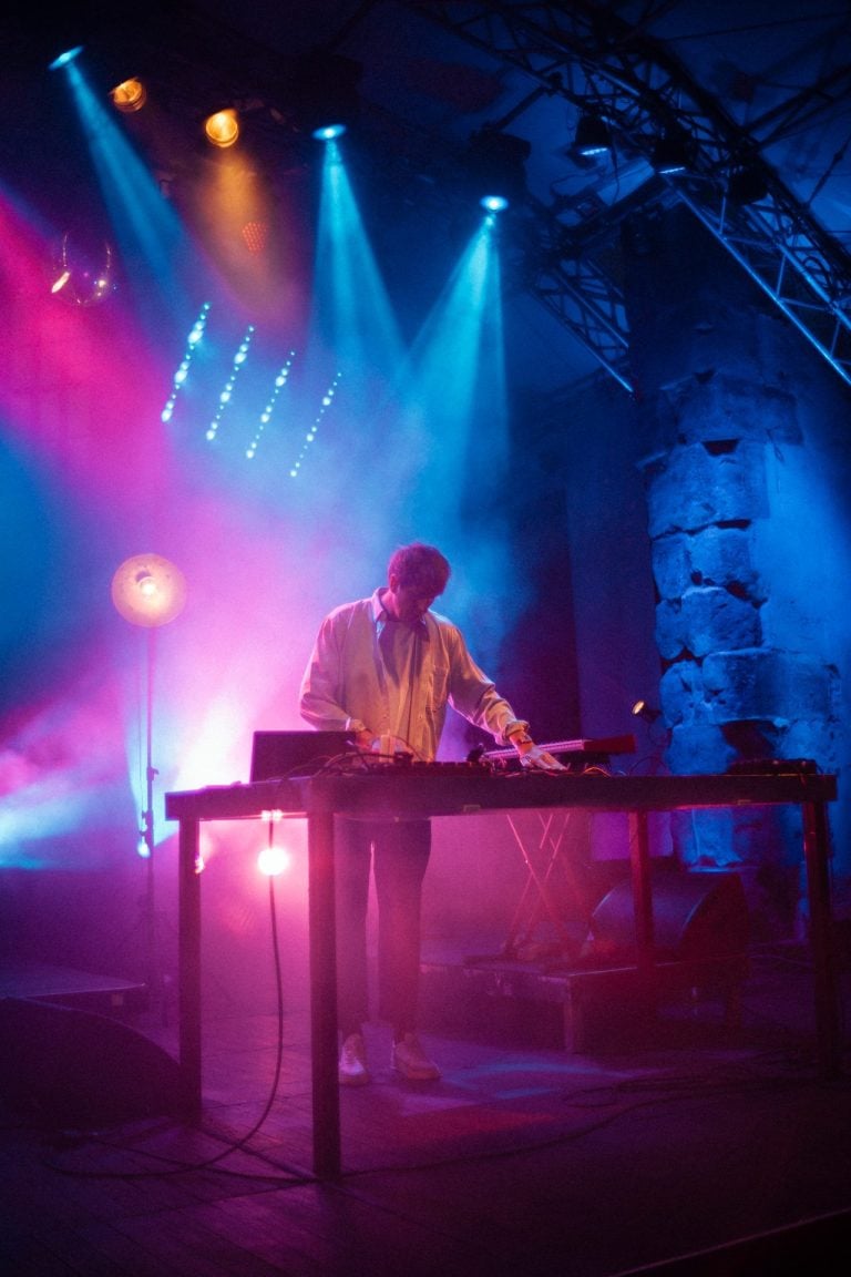 Musiker Christian Löffler steht hinter einem Tisch mit elektronischem Equipment auf einer Bühne, beleuchtet von atmosphärischem blauem und pinkem Scheinwerferlicht.