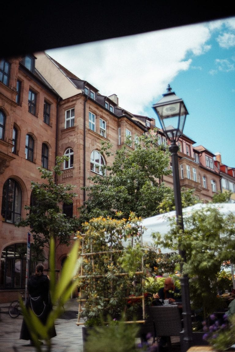 Ein historisches Sandsteingebäude in einer Stadt mit einer Straßenlaterne und einem Straßencafé im Vordergrund bei bewölktem Himmel.
