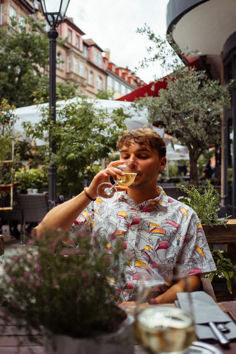 Ein junger Mann mit einem Tukan-Muster-Hemd trinkt entspannt ein Glas Weißwein in einem gemütlichen Straßencafé mit Pflanzen und Häuserfassaden.
