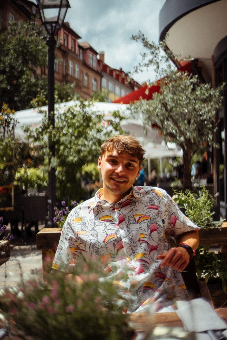 Ein junger Mann mit braunem Haar und Tukan-Muster-Hemd lächelt in einem sonnigen Straßencafé mit historischen Gebäuden im Hintergrund in die Kamera.