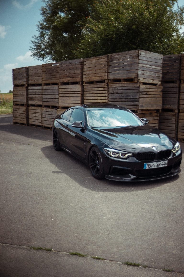 Ein schwarzes BMW Coupé steht auf einem asphaltierten Platz vor einer hohen Wand aus gestapelten, rustikalen Holzkisten unter einem sonnigen Himmel.