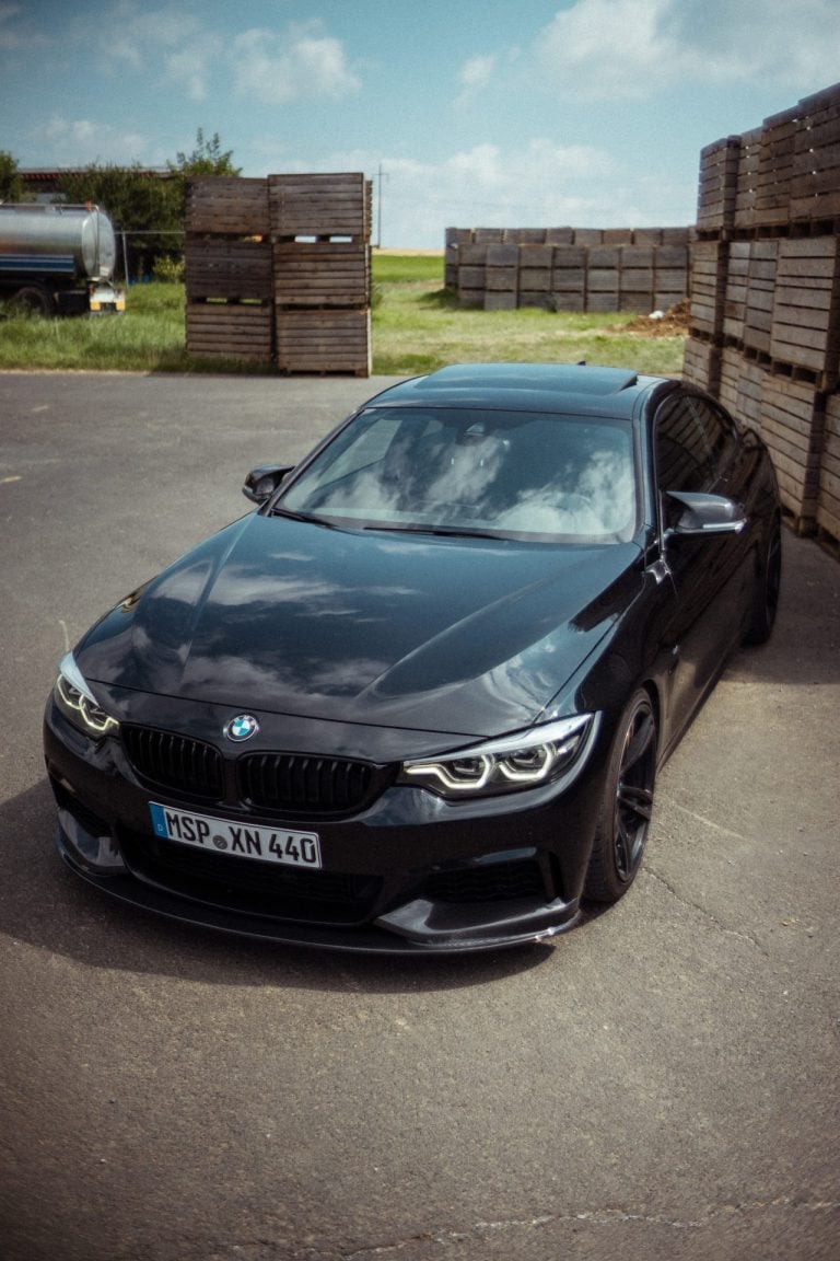 Ein schwarzes BMW Coupé steht auf einem asphaltierten Platz vor gestapelten Holzkisten an einem sonnigen Tag mit blauem Himmel und Wolken.