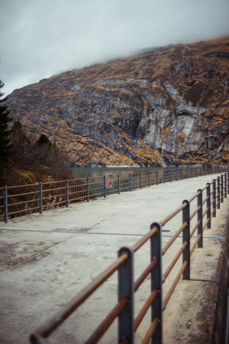 Ein betonierter Weg mit Metallgeländer führt an einem Stausee vorbei, im Hintergrund erheben sich steile, nebelverhangene Berge in herbstlichen Farben.