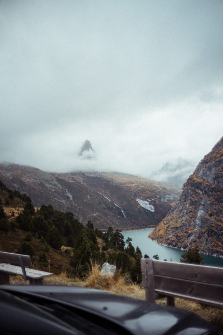 Blick von einer Anhöhe mit Holzbänken auf einen türkisfarbenen Bergsee, der von nebligen, wolkenverhangenen Bergen und Nadelwäldern umgeben ist.
