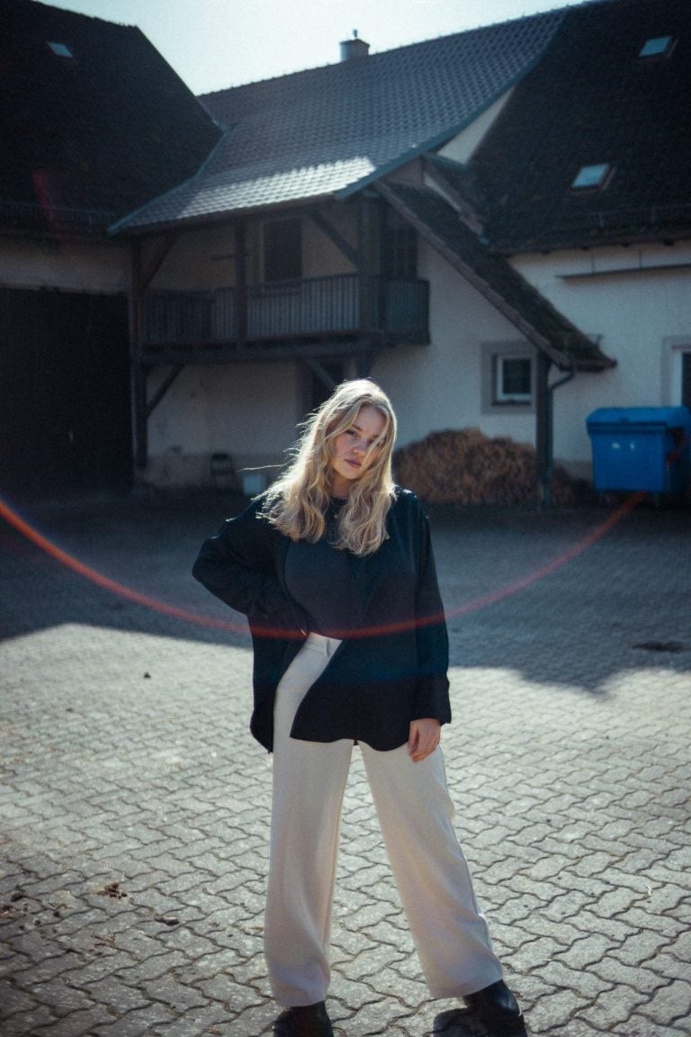 Eine junge Frau mit blondem Haar steht in einem schwarzen Hemd und heller Hose vor einem alten Bauernhaus auf einem gepflasterten Innenhof.
