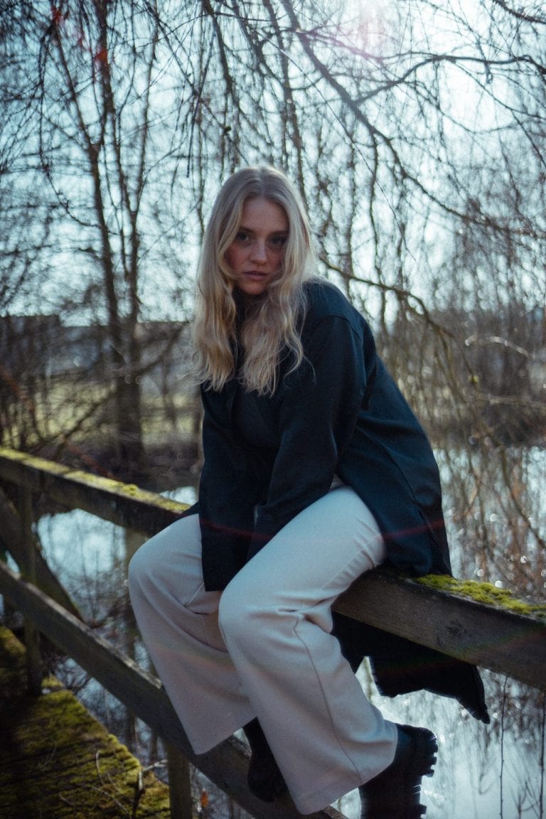 Eine junge Frau mit blondem Haar sitzt auf einem hölzernen Geländer vor einem Wald und einem See, bekleidet mit einem dunklen Hemd und heller Hose.