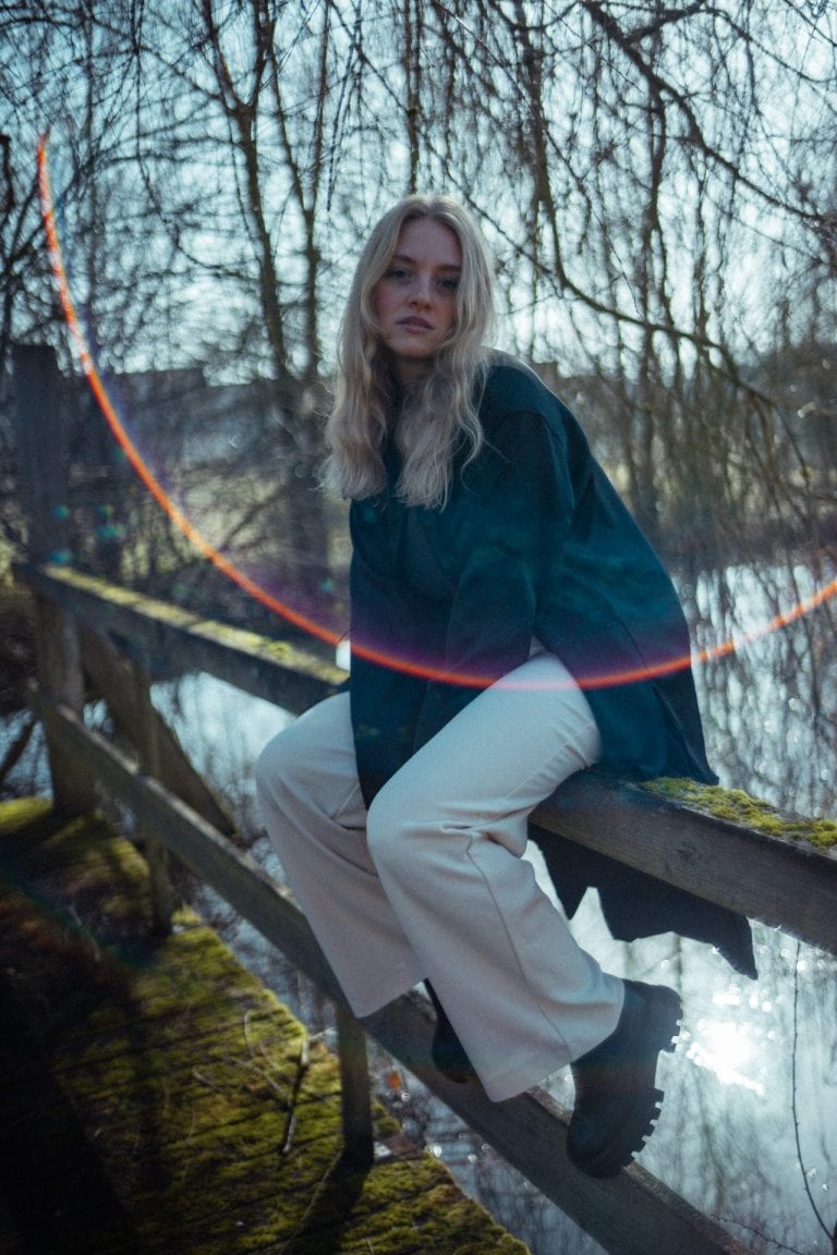 Eine junge Frau mit blondem Haar sitzt auf einem Holzgeländer an einem Fluss, während ein Lichteffekt eine bunte Kurve über das Bild zieht.