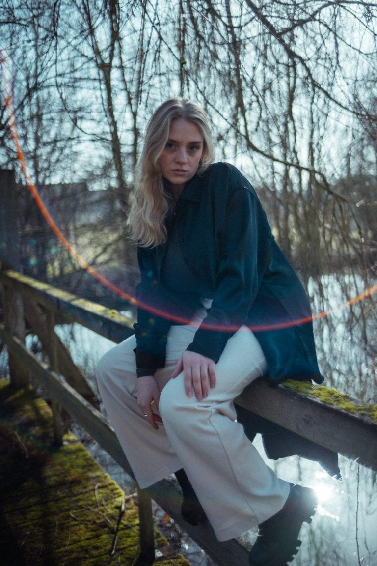 Eine junge Frau mit blondem Haar sitzt auf einem hölzernen Brückengeländer vor einer Waldkulisse mit einem Fluss im Hintergrund bei Tageslicht.